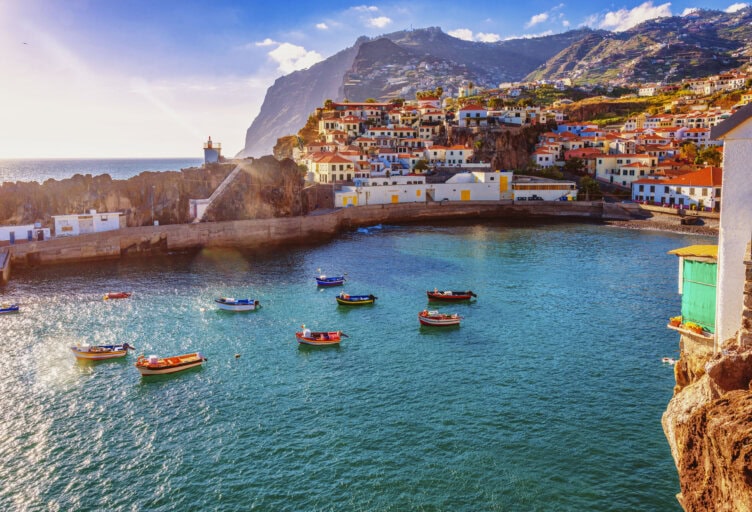 Colorful boats float in a coastal bay in Portugal, with distant mountains and a hillside village under a bright blue sky. The scenery is picture-perfect, making it clear why it's highlighted in every Portugal travel guide as the best time to visit.