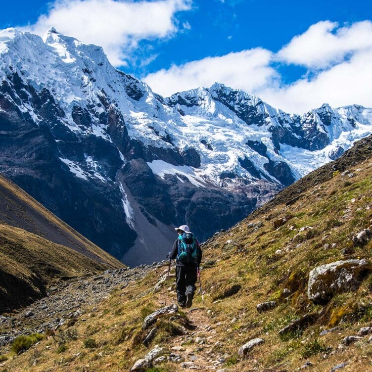 A solo traveler embarks on a journey along a rocky path in a mountainous landscape, surrounded by snow-capped peaks and set against a bright blue sky.
