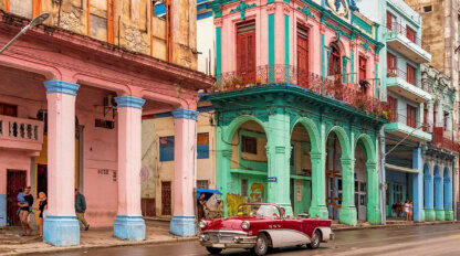 A classic car glides through the vibrant streets of Cuba, past colorful, weathered buildings with arches.