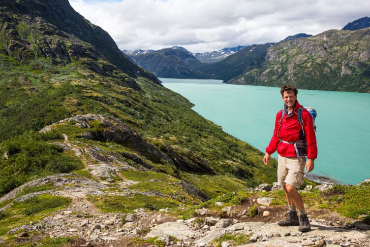 A person in a red jacket and shorts, choosing each step with care, hikes on a rocky trail overlooking a turquoise lake and mountains beneath a cloudy sky.