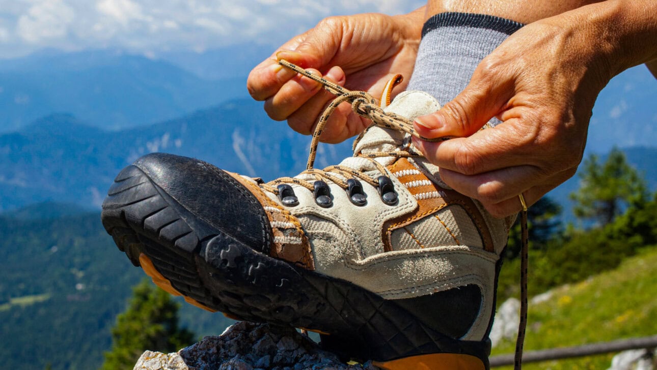 An adventurer expertly ties their hiking boots while sitting on a rock, surrounded by a majestic mountain landscape under a clear blue sky.