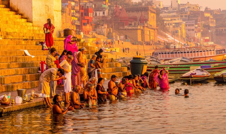 People are gathered at the riverbank steps in India, some bathing in the water, with colorful buildings as a vibrant backdrop. Boats are moored nearby, creating one of the top 15 things to do when exploring this lively destination.