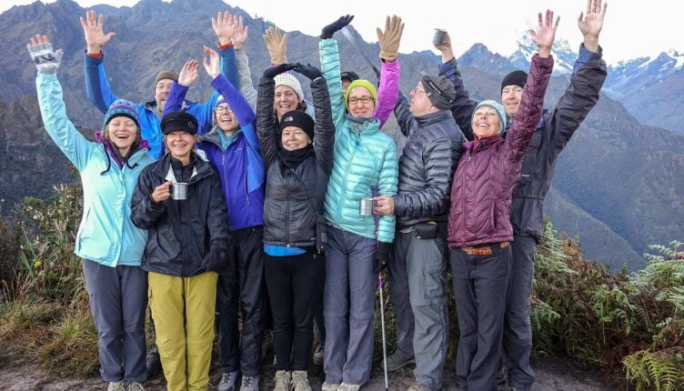 A group of people in hiking gear stand outdoors with arms raised, smiling. Mountains rise majestically in the background, reminiscent of an epic journey like hiking the Inca Trail.