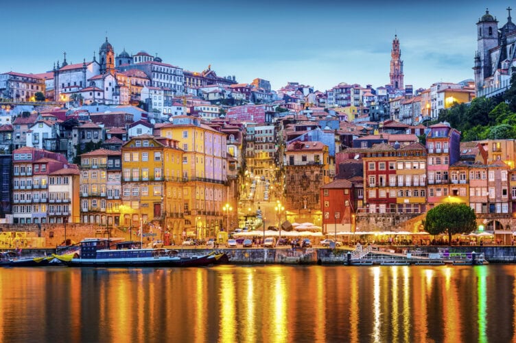 Discover the scenic beauty of Porto, Portugal, at sunset. Colorful buildings line the waterfront with reflections dancing on the Douro River. In the background, historic architecture and a church tower beckon travelers to explore this enchanting cityscape.