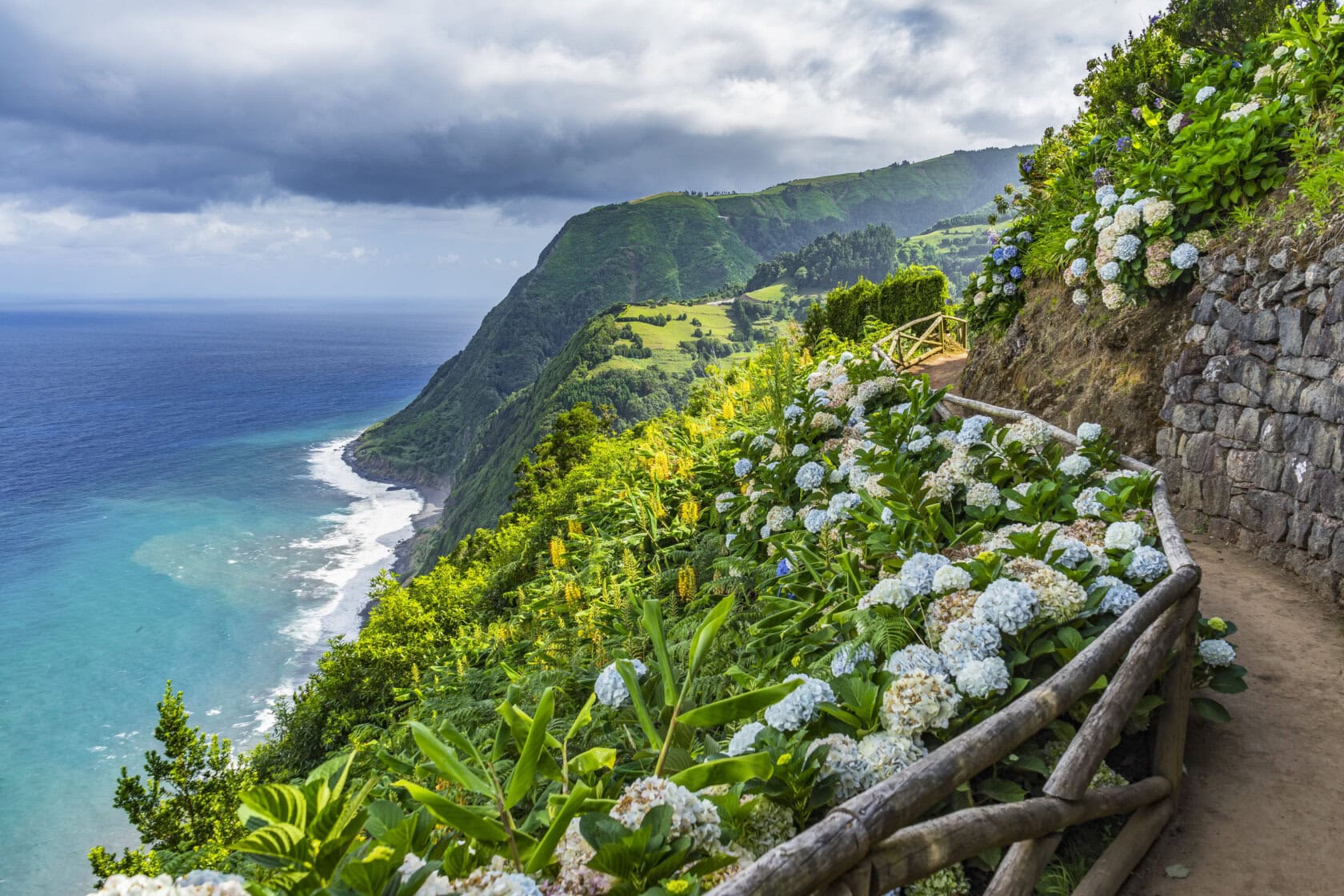A coastal cliff path in Portugal, bordered by blooming hydrangeas, overlooks a turquoise ocean and lush green landscape under a cloudy sky. Perfect for Portugal travel enthusiasts exploring the best time to visit this enchanting destination.