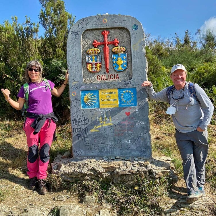 Two solo travelers stand beside a sign marking the Camino de Santiago in Galicia, Spain. The sign, adorned with graffiti and regional symbols, captures the spirit of their journey.