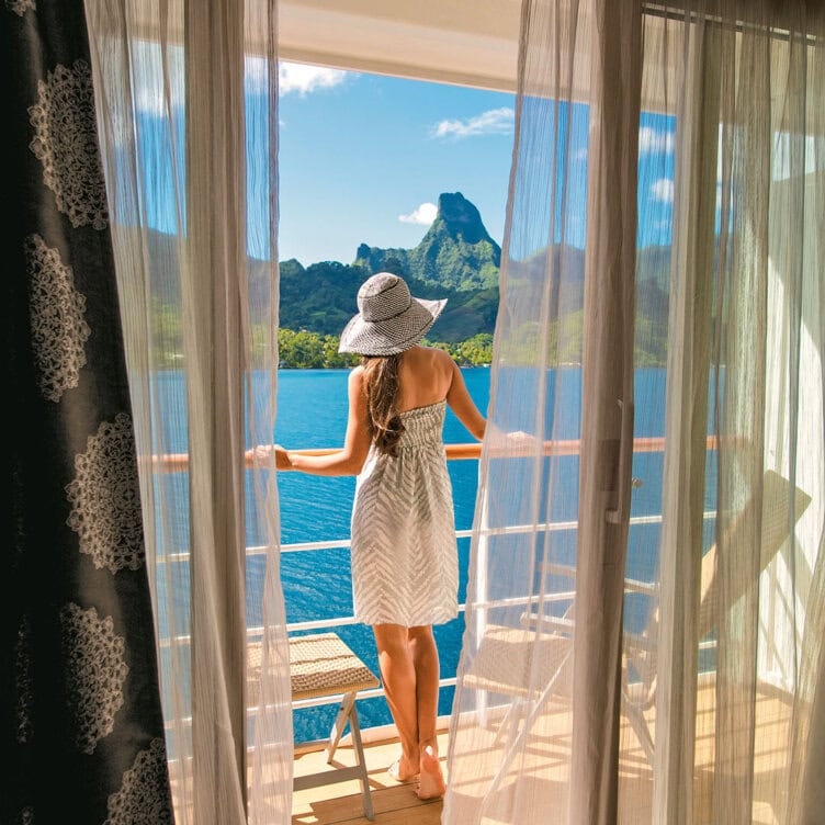 A solo traveler in a sun hat stands on a balcony, savoring the scenic ocean view with a mountain backdrop—an idyllic moment that defines unforgettable trips.