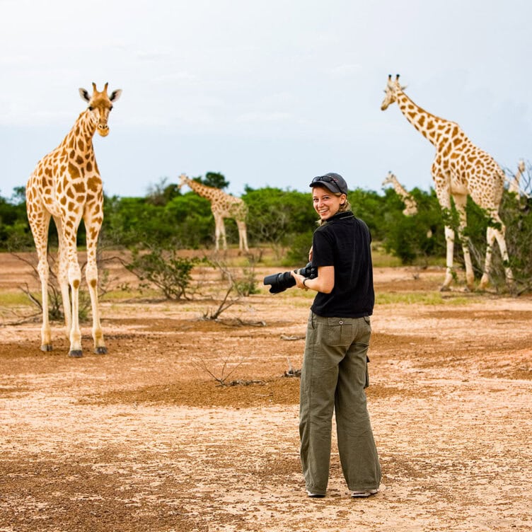 A solo traveler in a black shirt and cap stands in an open area, capturing the moment with their camera as several giraffes gracefully wander nearby.