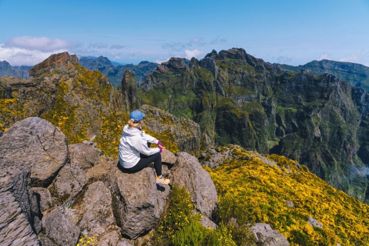 A person in a white jacket and blue cap sits on rocks, surrounded by yellow flowers, overlooking Portugal's stunning mountain landscape under a clear blue sky—an idyllic travel moment that captures the essence of adventure and tourism.