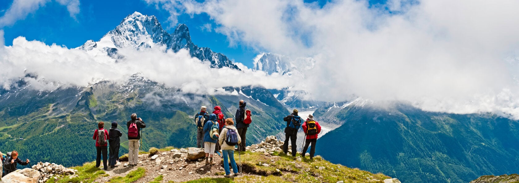A pack of hikers stands on a grassy hilltop, capturing photos of the majestic, snow-capped Alps partially veiled by clouds under a clear blue sky.