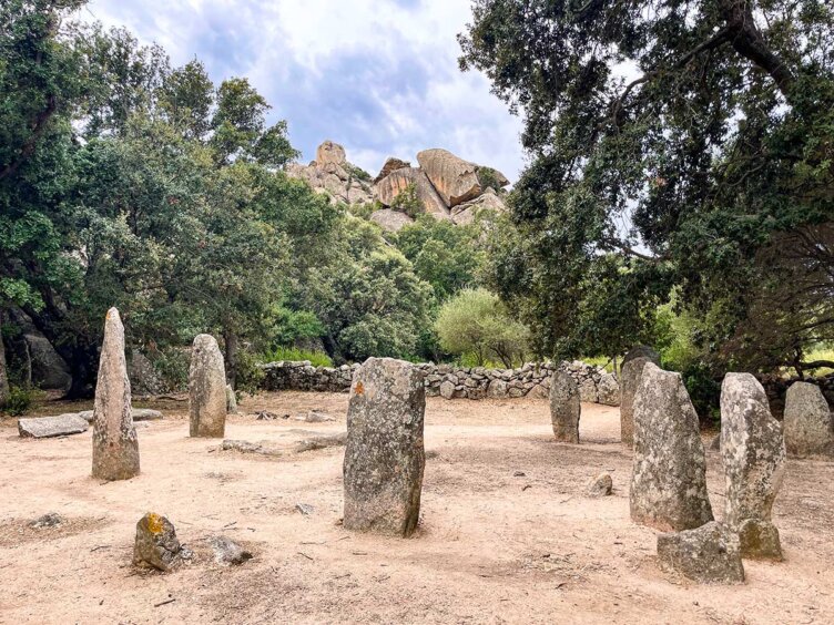 A stone circle surrounded by trees, with rugged Corsican hills in the background under a cloudy sky, inspires thoughts of hiking in Sardinia.