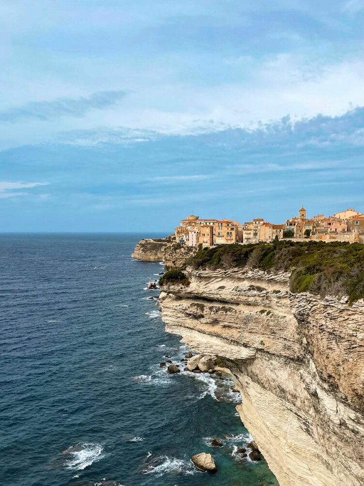 Set atop a stunning rocky coast, this cliffside town in Sardinia boasts beige buildings that gaze out over the blue sea under a partly cloudy sky, offering breathtaking views for hikers exploring the rugged terrain.