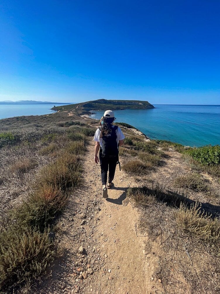A person with a backpack is hiking along a coastal trail in Sardinia, enjoying breathtaking ocean views on a clear day.