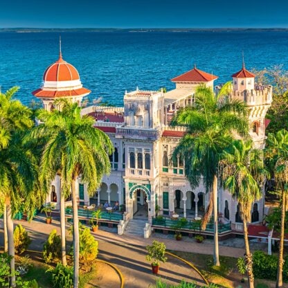 A grand historic building with a red-domed tower and ornate facade invites exploration, set among palm trees near the shimmering waters of Cuba under a clear blue sky.
