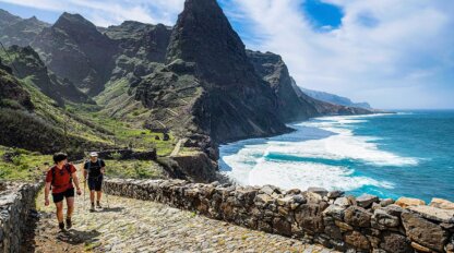 Two people explore a stone path along the mountainous Cape Verde coastline, with ocean waves crashing below under a partly cloudy sky.