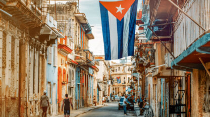 A large Cuban flag hangs over a narrow street lined with old buildings, inviting you to explore the vibrant culture as people stroll along the cobblestone road.