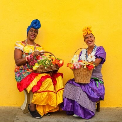 Two women in colorful dresses sit against a bright yellow wall, each holding a basket of flowers—a vibrant scene that feels like an invitation to explore the lively culture of Cuba.