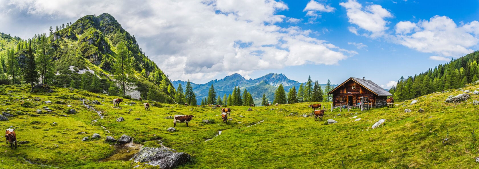 A wooden cabin sits on a grassy hillside with cows grazing, inviting the traveler to an adventure surrounded by trees and mountains under a partly cloudy sky.