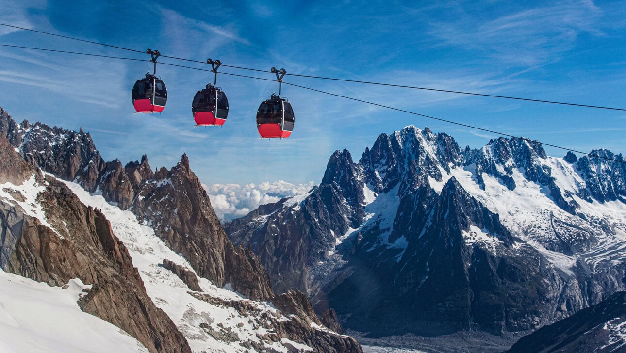 Three red cable cars travel over snowy mountains under a blue sky, offering a breathtaking view for any trip, with jagged peaks and a glacier visible in the background.