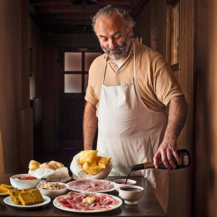 A traveler in an apron stands at a wooden table with various plates of cured meats, bread, and small bowls, pouring a dark liquid into a cup—fueling up for his next adventure.