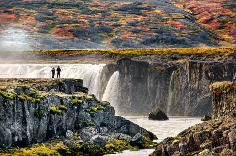 Two people stand near the edge of rocky cliffs after a scenic hike, overlooking a large waterfall with colorful autumn foliage covering the surrounding hills.