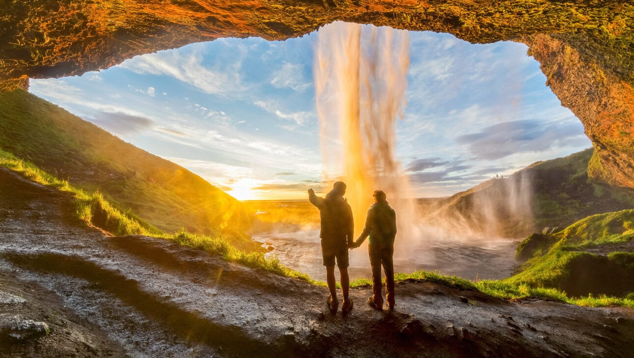 Two people stand in a cave behind a waterfall, holding hands and facing the sunset over a grassy landscape, their trip guided by the promise of adventure.