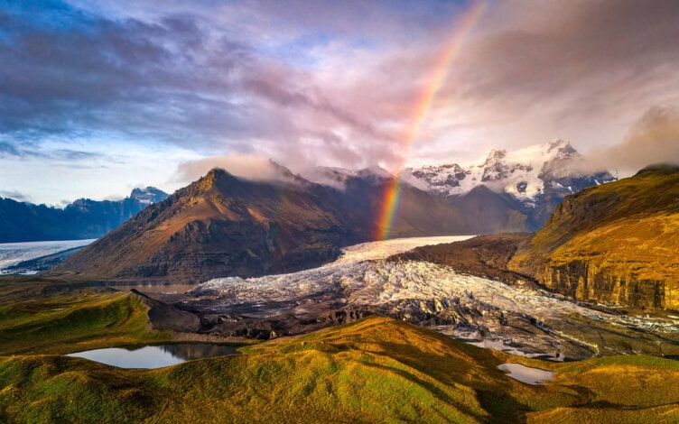 A glacier winds through green hills under a cloudy sky, inviting the adventurous traveler to hike beneath a rainbow arching over snow-capped mountains in the background.