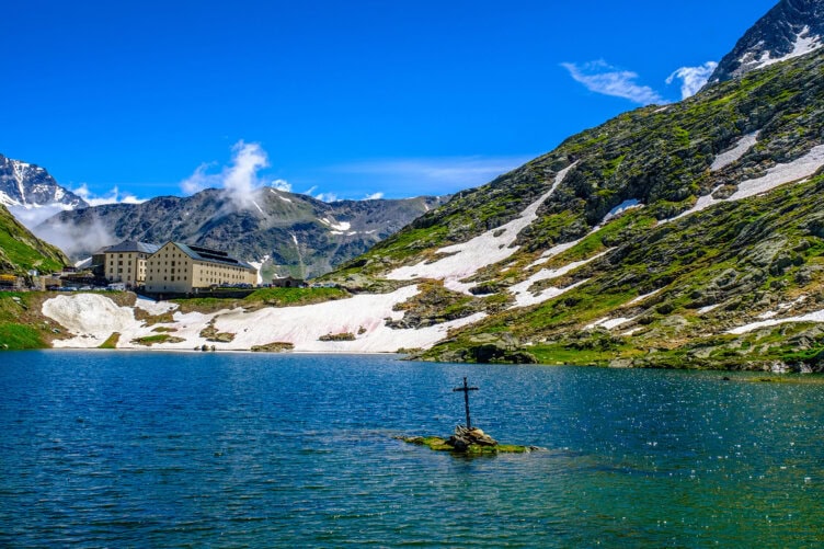 A large alpine lake with a cross on a small rock in the water, surrounded by mountains and patches of snow, invites adventure seekers. A building on the shore under a blue sky hints at the perfect base for your guided mountain tour.