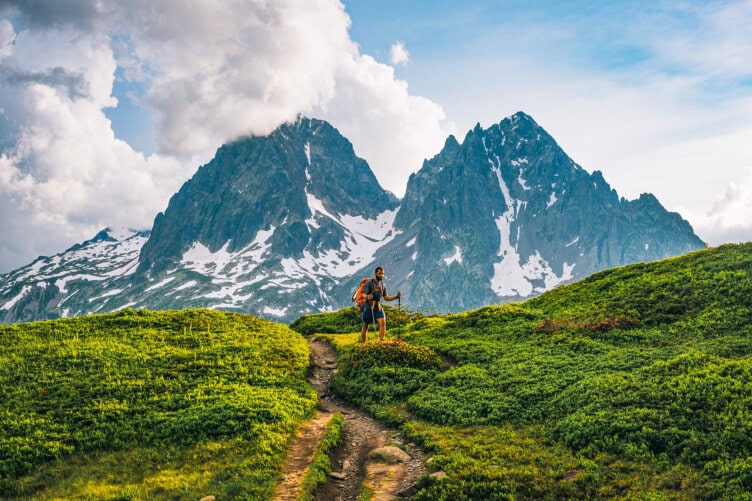 An adventurous hiker with a backpack and trekking poles walks along a grassy trail towards tall, snow-capped mountains under a partly cloudy sky.