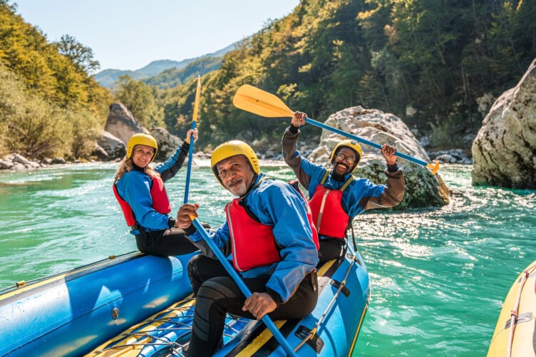 Three adventure seekers in life jackets and helmets paddle a blue inflatable raft down a river surrounded by trees and rocks on a sunny day, embracing the thrill of outdoor travel.