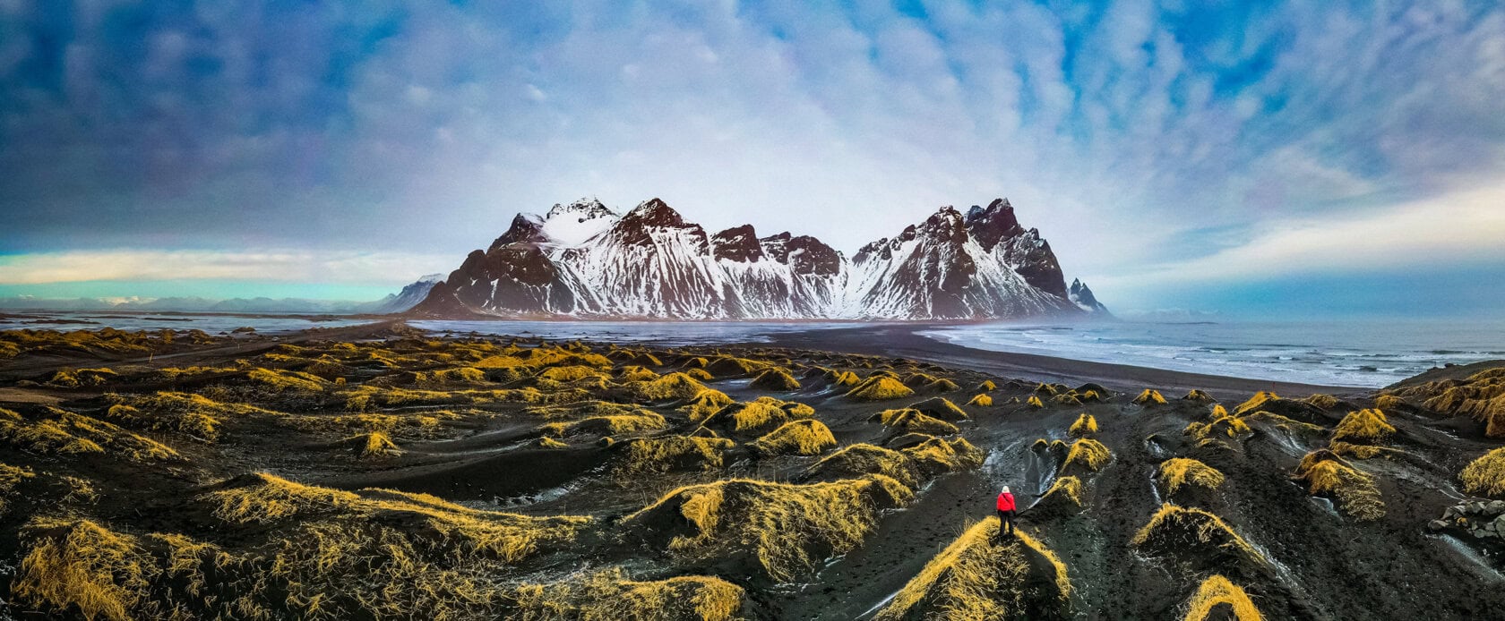 A person in a red jacket stands among grassy sand dunes, ready for a hike, with snow-capped mountains and a cloudy sky in the background.