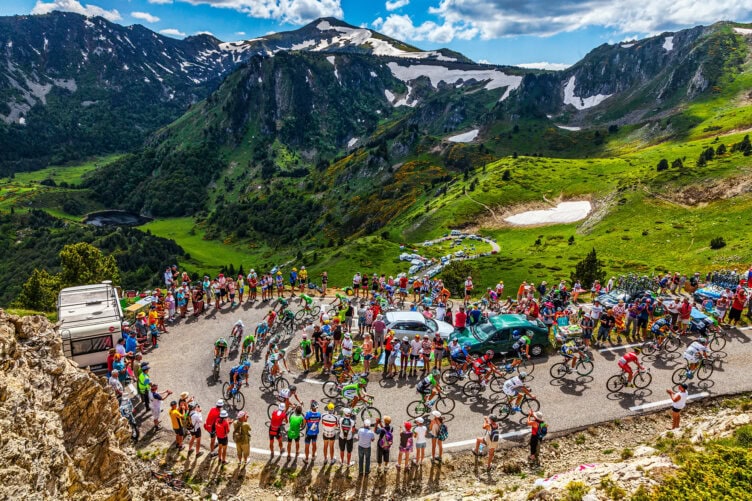 Cyclists ride up a winding mountain road, reminiscent of a tour guided through scenic terrain, lined with spectators and vehicles, with green hills and snow-capped mountains under a partly cloudy sky.