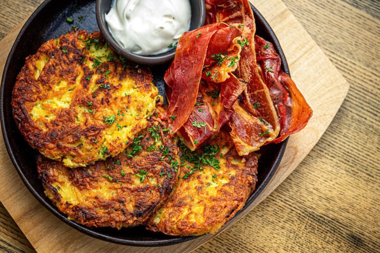 A perfect meal after a hike: three golden-brown potato pancakes, crispy strips of cured meat, a small bowl of sour cream, and a garnish of chopped herbs on a wooden board.
