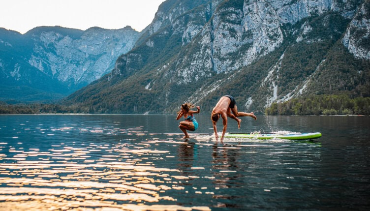 Two people fall off a paddleboard into a calm mountain lake, beginning an unexpected adventure surrounded by forested hills and rocky peaks—a perfect spot after a scenic hike.
