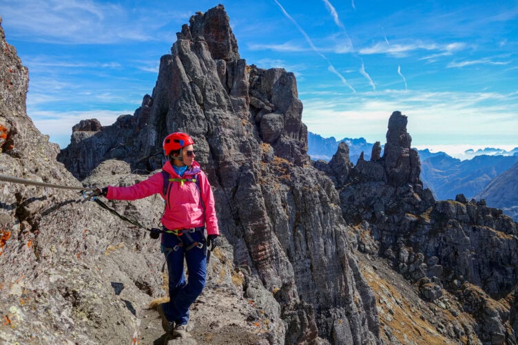A climber wearing a helmet and harness stands on a rocky mountain ledge, holding onto a safety cable, as a guide leads the trip among jagged peaks beneath a blue sky.
