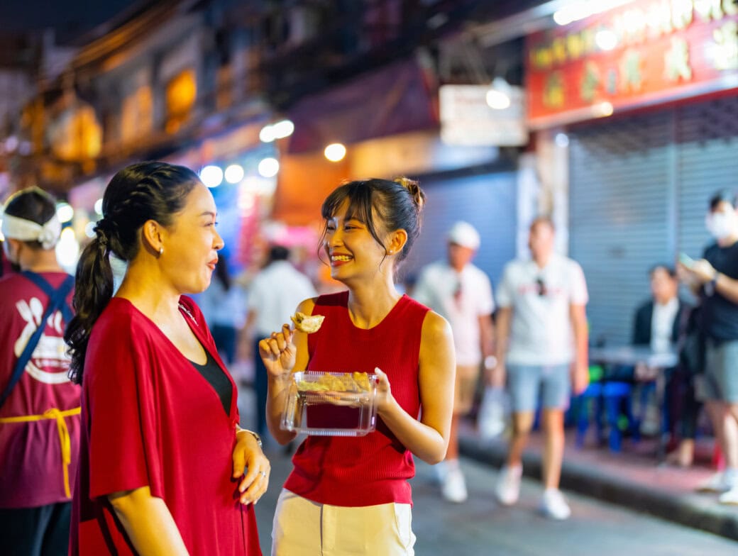 Two women talk and smile while eating street food at a busy outdoor market at night—an authentic Bangkok travel experience, with other people and vibrant food stalls in the background.