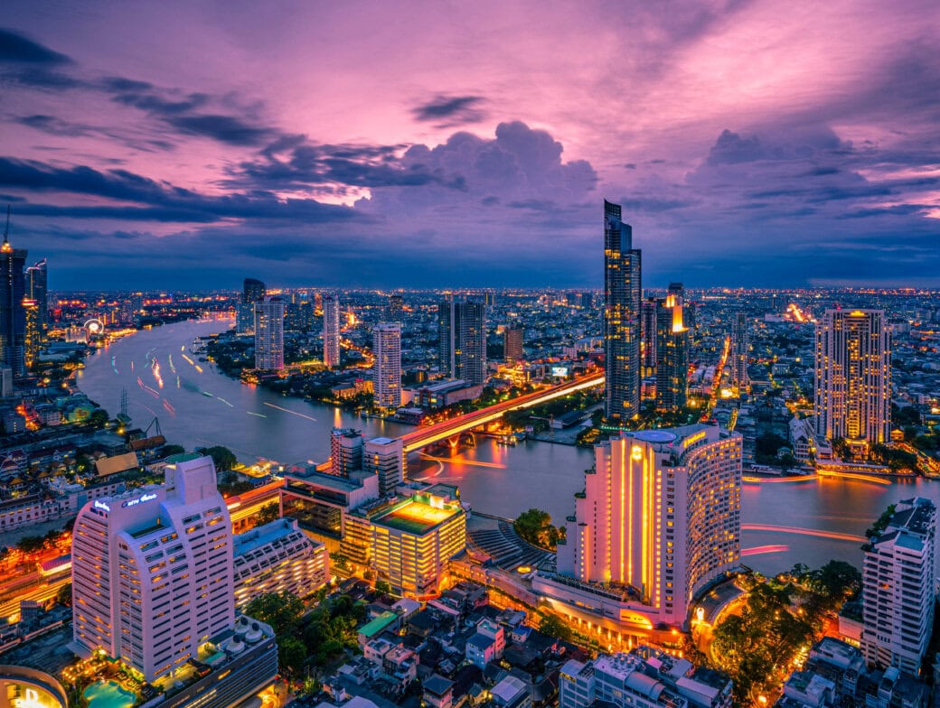 A city skyline at dusk with illuminated buildings and a river running through the center under a dramatic, cloudy sky, capturing the essence of Bangkok attractions and the vibrant spirit of Bangkok travel.