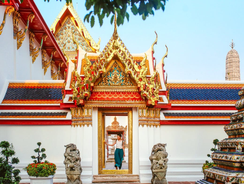 A person enters an ornate temple in Bangkok, adorned with red, gold, and white details, flanked by two stone statues—a must-see for those seeking unique things to do while they travel.