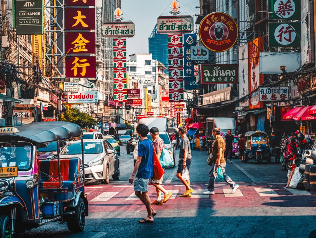 People cross a busy street filled with cars, tuk-tuks, and motorcycles in an urban area lined with buildings and storefronts displaying signs in Thai script—a classic scene for those seeking authentic Bangkok travel experiences.
