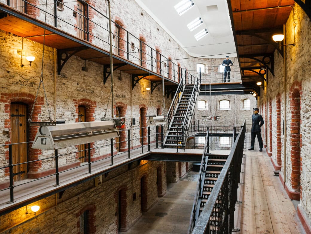 Interior view of an old multi-level prison in Cork with stone walls, metal railings, and staircases; two uniformed guards are visible on different levels—one of the intriguing Cork attractions for history enthusiasts.