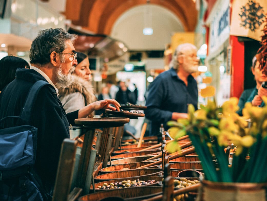 People browse and select food items from wooden barrels at an indoor market in Cork, with flowers and crowds in the background—a must-see among the top 10 Cork experiences and things to do in Cork.