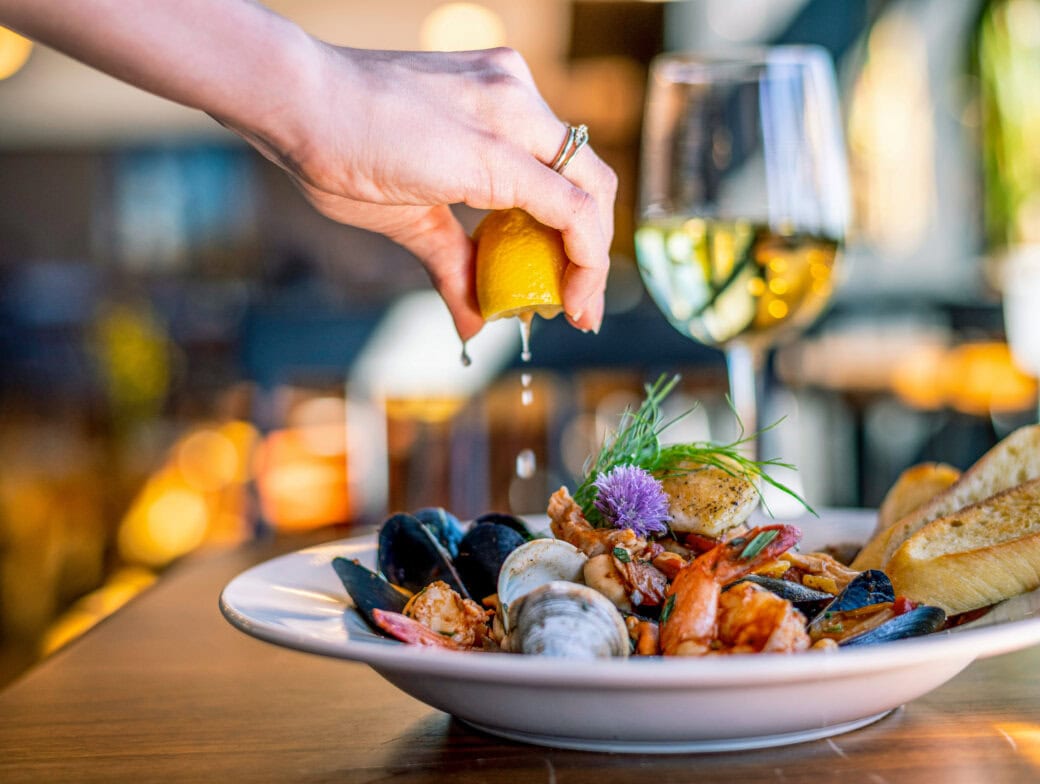 A hand squeezes lemon over a seafood dish with mussels, clams, shrimp, and bread, with a glass of white wine in the background—perfect for experiencing Cork tourism and savoring local flavors after exploring top Things to Do in Cork.
