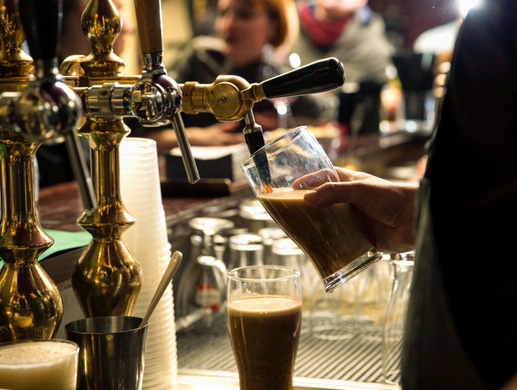 A bartender in Cork pours a dark beer from a tap into a glass at a bar, with another filled glass and empty cups on the counter—perfect for those seeking top 10 things to do in the city.
