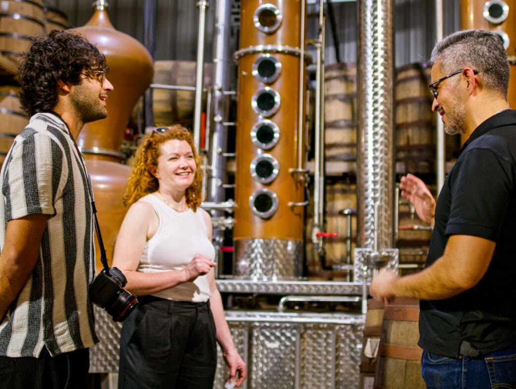 Three people stand and talk inside a distillery surrounded by barrels and large distillation equipment; one person holds a camera, capturing one of the top things to do in Cork for those interested in unique Cork attractions.