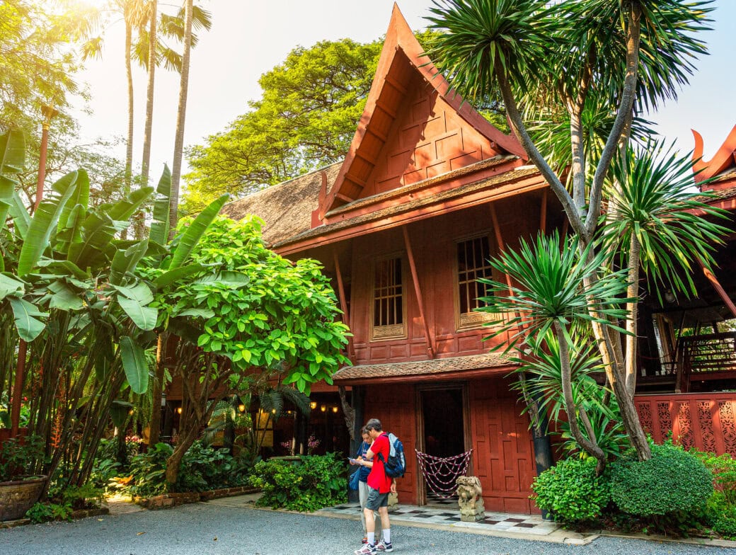 A person with a red backpack stands outside a traditional wooden Thai house surrounded by lush tropical plants and trees, capturing the charm of Bangkok attractions and unique things to do in Bangkok.