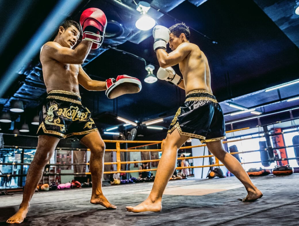 Two men spar in a boxing ring, wearing gloves and traditional shorts, inside a gym filled with training equipment—an exciting glimpse into Bangkok activities and one of the top things to do in Bangkok for sports enthusiasts.
