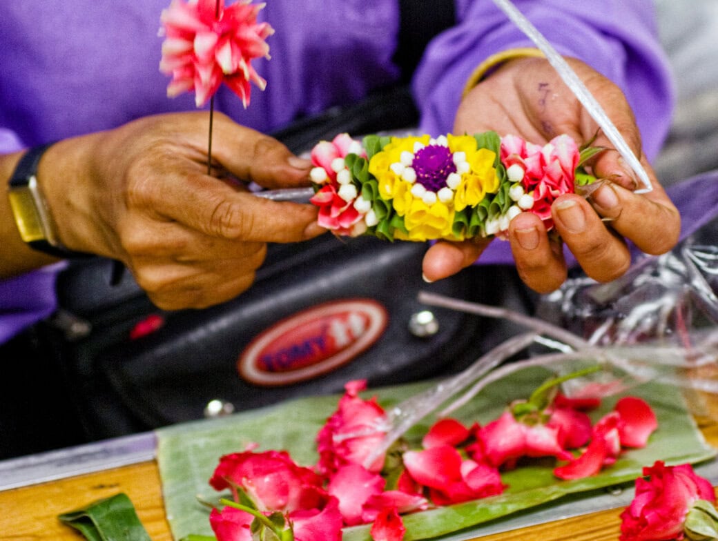 A person arranges colorful artificial flowers into a garland, with pink rose petals and green leaves on a table—a perfect glimpse of the creative crafts you can discover among Bangkok attractions.