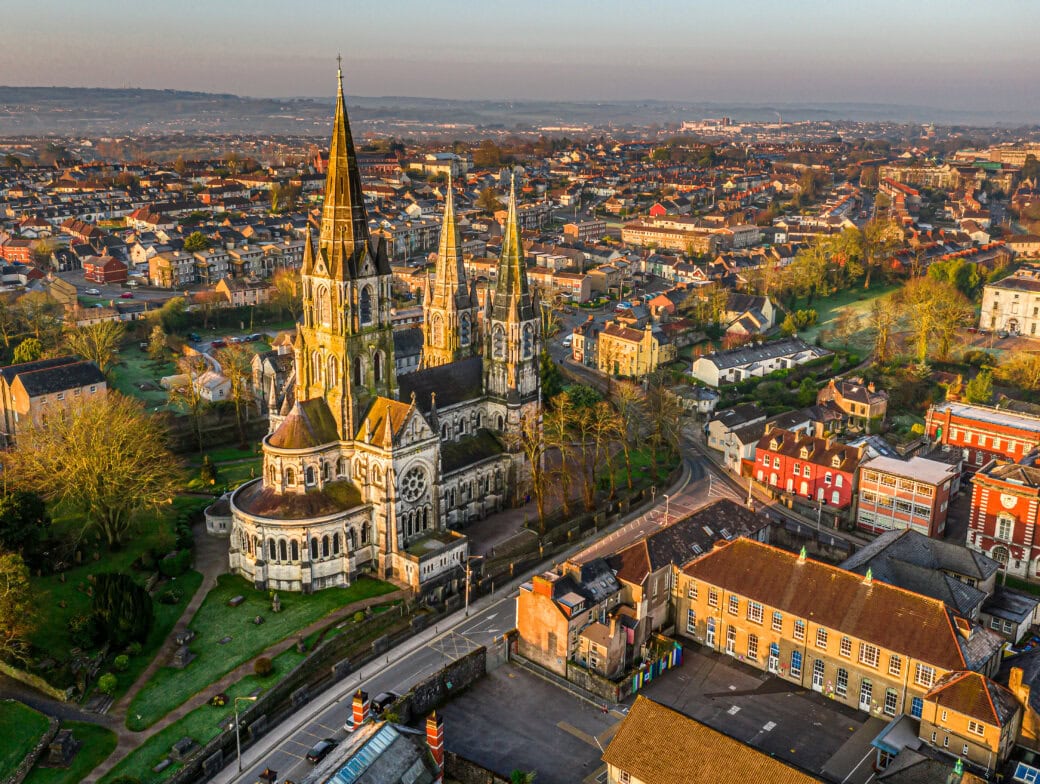 Aerial view of a large Gothic-style cathedral surrounded by trees and buildings in a cityscape at sunrise, highlighting one of the must-see Cork attractions for those exploring Cork tourism.