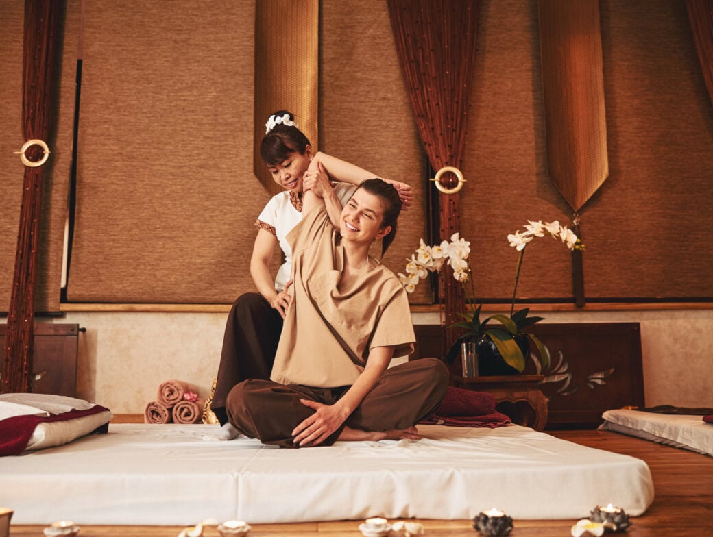 A massage therapist performs a shoulder stretch on a seated client inside a spa in Bangkok, surrounded by candles, towels, and potted orchids—one of the relaxing things to do while you travel.