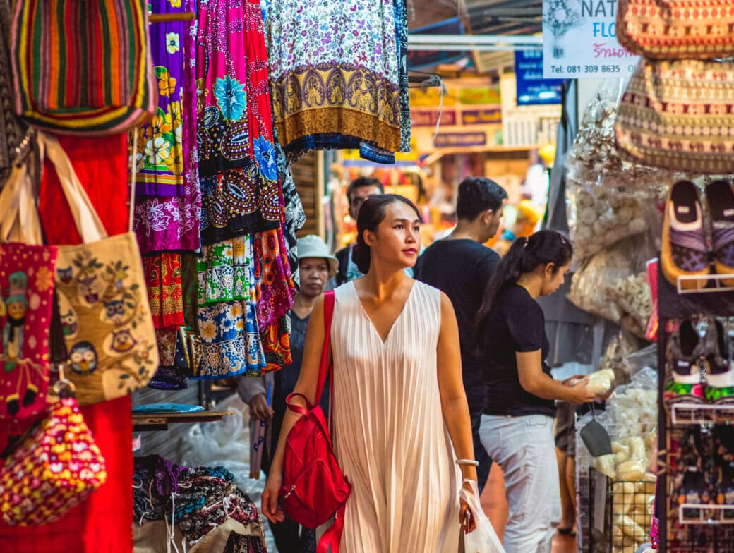 A woman in a white dress walks through a colorful, busy market with hanging clothes, bags, and shoppers—a perfect snapshot of Bangkok attractions for anyone exploring top things to do in Bangkok.
